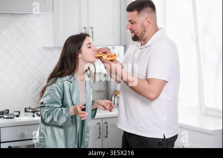 Glückliches verheiratetes Paar mit romantischem Pizza Date Night zu Hause, Mann füttert Frau mit Stück Pizza. Ehe- und Pflegekonzept, Teamwork in der Küche, Stockfoto