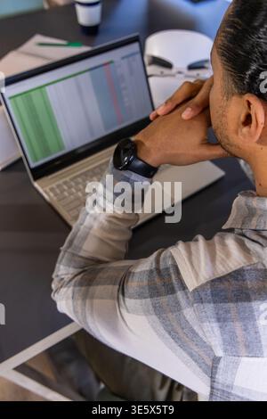 Mann im karierten Hemd, der sich nach vorne lehnt, der Tisch auf dem Laptop am Schreibtisch mit Maske, Uhr, Kaffee überprüft Stockfoto