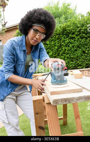 Frau, die Holzplanke auf Sägepferd im Garten mit schnurgebundenem Schleifgerät schleift, mit Brille Denim-Hemd Stockfoto