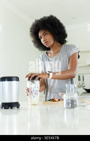 Afrikanische Frau, die an der Küchenzeile steht und einen klaren Becher hält und den Deckel neben dem Mixersockel hält Stockfoto