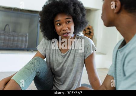 Afroamerikanische Freunde sitzen auf dem Boden in der Nähe von Kamin und Brennholz und unterhalten sich in Workout-Kleidung Stockfoto