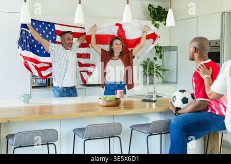 Gruppe von vier Freunden, die auf der Kücheninsel mit Fußball und rotem Trikot anfeuern und Fahnen schwenken Stockfoto