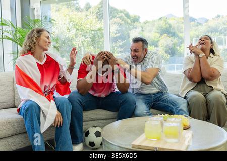Verschiedene Freunde reagieren auf Fußballspiele im Wohnzimmer auf einem Sofa mit Fußball, roten Trikots Stockfoto