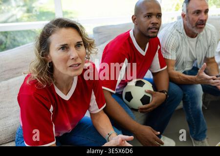 Verschiedene Freunde, die Fußballspiele auf einem leichten Sofa beobachten, in roten Trikots mit Ball, Kopierraum Stockfoto