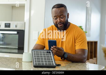 Mittelalter afroamerikanischer Mann mit blauem Smartphone neben tragbarem Solarpanel auf der Küchentheke Stockfoto