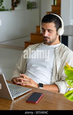 Mann sitzt zu Hause am Holztisch und trägt Kopfhörer und beigefarbenes Overshirt, Laptop in der Nähe Stockfoto