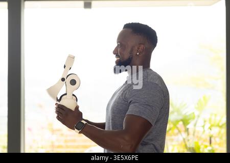 Afrikanischer Mann, der in einem hellen Raum in der Nähe von Glas steht, weißes Headset hält, graues T-Stück trägt, Kopierraum Stockfoto