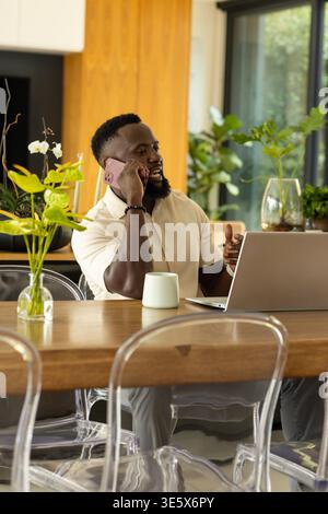 Mann am Heimtisch mit Kurzarm-Hemd, Anruf auf rosa Telefon und Arbeit am Laptop Stockfoto
