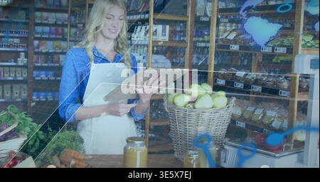 Ein Angestellter in einer Hemdschürze aus Denim, der den Lagerbestand in einem kleinen Geschäft mit Tablettengläsern aus Clipboard überprüft Stockfoto