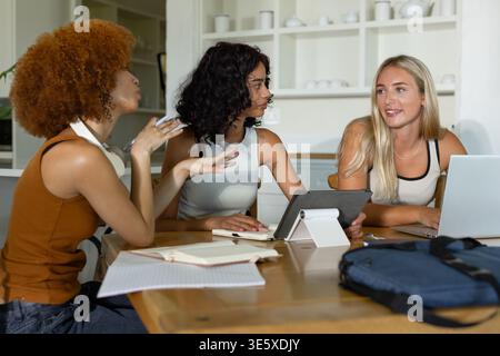 Drei Freundinnen sitzen am Küchentisch und sprechen über Notizen mit Laptop, Tablet auf weißem Ständer Stockfoto
