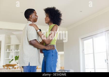 Afroamerikanisches Paar in 20ern, das zu Hause steht und sich umhüllt und Smartwatch in der Nähe von Glastüren zeigt Stockfoto