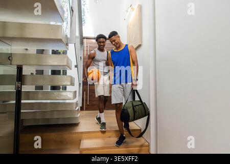 Verschiedene männliche Freunde laufen, die Holztreppen im Flur hinuntergehen, mit Basketball und Seesack Stockfoto