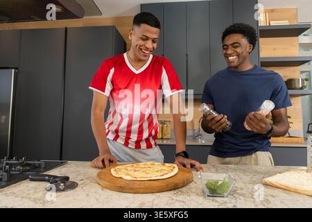 Lächelnde männliche Freunde bereiten Pizza auf Marmorinsel in moderner Küche mit Pizzaschneider zu Stockfoto