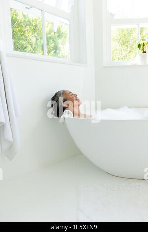 Hochrangige afroamerikanische Frau, die in der Badewanne liegt und den Kopf auf einem gerollten Handtuch in Schaum an den Fenstern ruht Stockfoto
