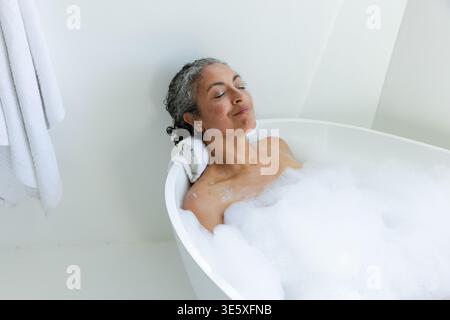 Ältere afroamerikanische Frau, die sich in einer freistehenden Wanne zurücklehnt, sich zwischen Seifenblasen entspannt, Handtuchkopfstütze Stockfoto