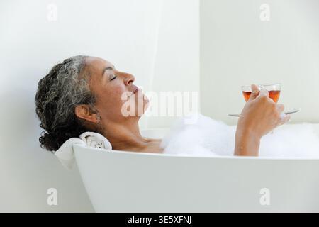 Ältere afroamerikanische Frau, die in der Badewanne liegend war, mit Blasen im Badezimmer, mit Glasbecher Stockfoto