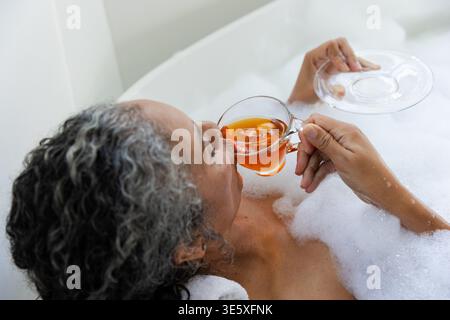 Badfüllung, Seniorin, die sich in Schaum zurücklehnt, Glas-Teetasse und Untertasse hält, Ruhe sucht Stockfoto