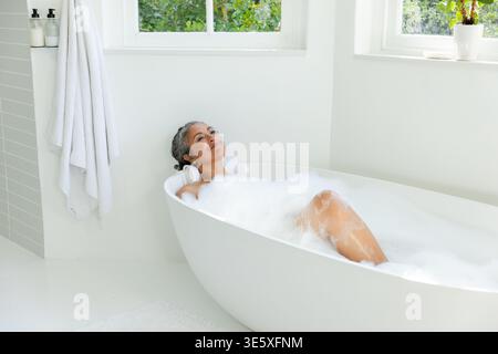 Ältere afroamerikanische Frau, die in einem hellen Badezimmer in einer freistehenden Badewanne mit Schaum liegt Stockfoto