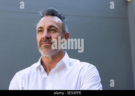 Mann mittleren Alters, der an einer dunkelgrauen Wand mit horizontaler Naht steht und ein weißes Hemd mit Knopfverschluss trägt Stockfoto