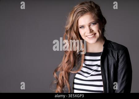 Frau mit langen Haaren und schwarz-weiß gestreiftem Hemd lächelt vor der Kamera. Sie trägt eine Lederjacke und ist glücklich und selbstbewusst Stockfoto