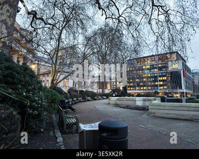 London, UK - 02. Februar 2026: Ruhiger Abend im Trinity Square Gardens London, UK Park mit Bänken, blattlosen Bäumen und beleuchteten Bürogebäuden. Stockfoto