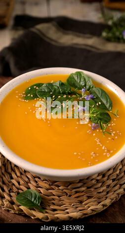 Ein lebhaftes orangenes Gemüsepüree, serviert in einer Keramikschale, garniert mit Babyspinatblättern und Sesamsamen. Die rustikale Szene bietet natürliches Licht Stockfoto