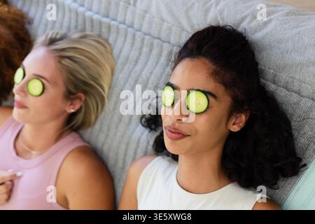 Verschiedene Freundinnen, die Tanktops tragen, liegen zu Hause auf einer gerippten grauen Decke mit Gurkenscheiben Stockfoto