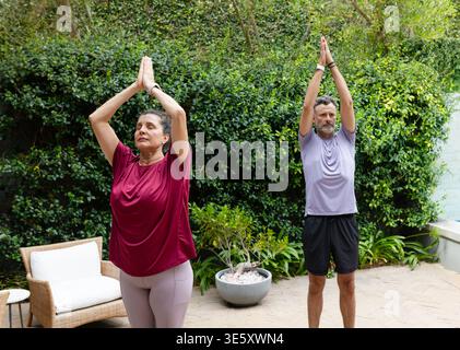 Seniorenpaar üben Yoga im Stehen auf der Terrasse in Aktivkleidung mit Korbstuhl, Pflanzgefäß, Uhren Stockfoto