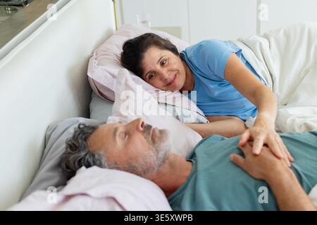 Verschiedene Paare, die zu Hause auf dem Bett liegen und T-Shirts tragen, eine Frau, die über die Brust greift Stockfoto