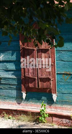 Alte geschlossene Holzfenstern mit abblätternder brauner Farbe an einer blau verwitterten Holzhauswand. Nahaufnahme. Stockfoto