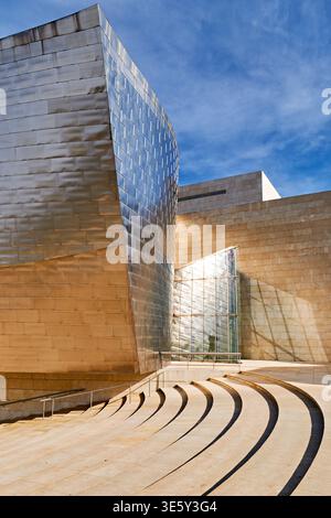 Besucher erkunden die markanten Formen und Materialien des Guggenheim Museums in Bilbao, Spanien an einem klaren Tag mit blauem Himmel. Stockfoto