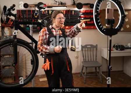Eine Fahrradmechanikerin, die ein Tutorial über Kamera und Vlogging für Abonnenten in ihrer Fahrradwerkstatt aufnimmt, indem sie ein Smartphone zum Bloggen benutzt. Stockfoto