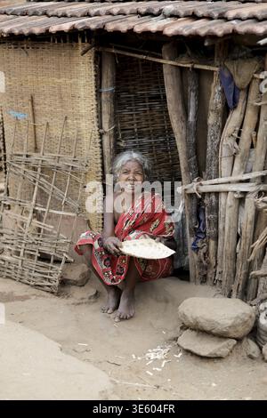 Odisha, Indien - 26. März 2026: Blick auf eine lächelnde Frau in einem roten Saree, die am rustikalen Eingang eines strohgedeckten Hauses hockt und ein einfaches Leben in sich widerspiegelt Stockfoto