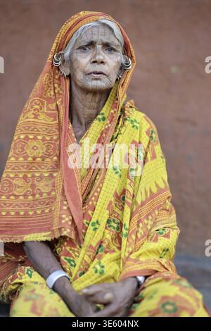 Odisha, Indien - 26. März 2026: Blick auf eine ältere Frau mit verwittertem Gesicht, geschmückt mit einem leuchtenden gelb-orangen Sari, die feierlich an einem sitzt Stockfoto