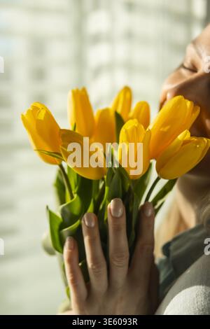 Frau, die einen frischen Strauß gelber Tulpen hält, sie mit geschlossenen Augen nahe an ihr Gesicht bringt, ihren Duft genießt und die Freude des Spri feiert Stockfoto