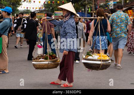 Hoi an, Vietnam – Februar 2026: Straßenverkäufer mit Obstkörben und Schulterpfosten spazieren durch die geschäftige Marktstraße Stockfoto
