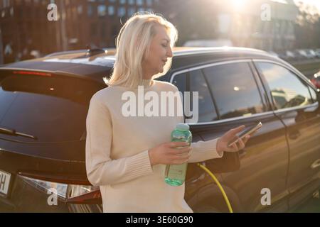 Umweltbewusste blonde Frau, die während des Ladevorgangs neben einem Elektrofahrzeug steht. Stockfoto