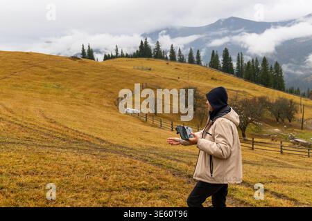 Drohnenfahrer in Berglandschaft. Person, die eine Drohne auf einem grasbewachsenen Hügel mit Zäunen, Bäumen und nebeligen Bergen unter einer bewölkten Oberfläche fliegt Stockfoto
