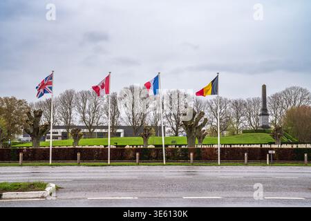Straßenblick auf den Friedhof Essex Farm in Ypern, Belgien. Flaggen aus Großbritannien, Kanada, Frankreich und Belgien fliegen vor dem 49th West Riding Division Memorial Stockfoto