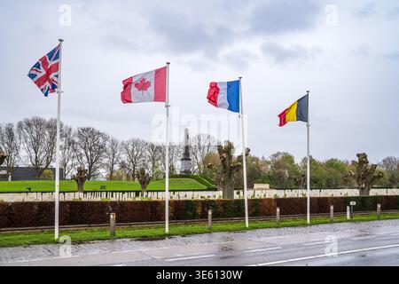 Straßenblick auf den Friedhof Essex Farm in Ypern, Belgien. Flaggen aus Großbritannien, Kanada, Frankreich und Belgien fliegen vor dem 49th West Riding Division Memorial Stockfoto