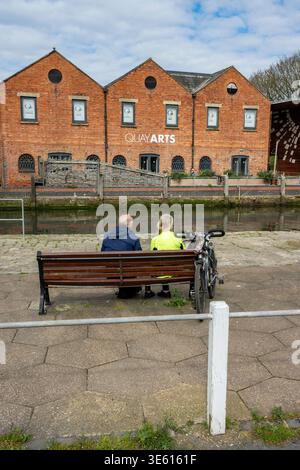 Älteres Ehepaar, das auf einer Bank sitzt, während es auf einer Radtour gegenüber dem Quay Arts Centre an der Flussmedina in Newport, Isle of Wight, England, Großbritannien, fährt Stockfoto