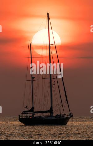 Silhouette einer Segelyacht mit Mast, ausgerichtet gegen die große untergehende Sonne in Phuket, Thailand Stockfoto