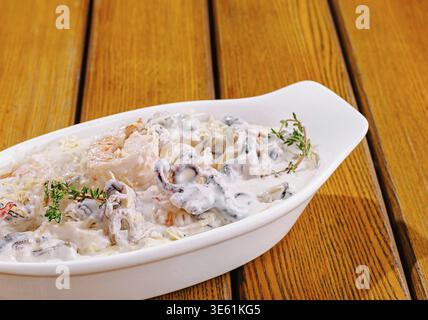 Essen, Ein weißes Gericht gefüllt mit Garnelen und Pilzen in einer cremigen Sauce, sitzt auf einem rustikalen Holztisch. Frische Kräuter garnieren das Gericht und betonen einen Feinkost Stockfoto