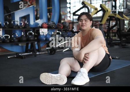 Übergewichtiger Mann, der nach dem Training im Fitnessstudio auf einer Matte sitzt. Konzept der Fitness-Reise, Körperpositivität, Erholung und gesunder Lebensstil Stockfoto