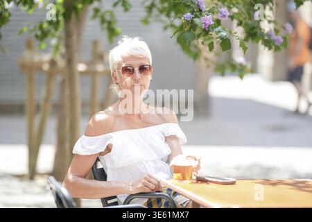 Eine Frau in einem weißen Hemd sitzt mit einer Tasse Kaffee an einem Tisch. Sie trägt eine Sonnenbrille und einen Hut Stockfoto