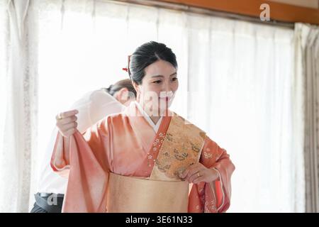 Eine Frau in einem traditionellen japanischen Kimono passt ihr Outfit an. Die Szene spielt in einem Raum mit Fenster, und hinter ihr steht ein Mann. Th Stockfoto