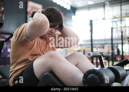 Übergewichtiger Mann, der im Fitnessstudio Sit-ups macht, schwitzt und nach dem Training müde aussieht. Konzept von Fitness, Gewichtsverlust Reise, Körperpositivität, DE Stockfoto