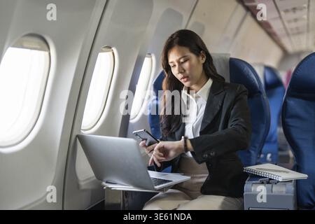 Eine professionelle Frau, die ihren Zeitplan auf einem Smartphone verfolgt, während sie während eines Fluges an ihrem Laptop arbeitet, wobei sie den Schwerpunkt auf Zeitmanagement legt Stockfoto