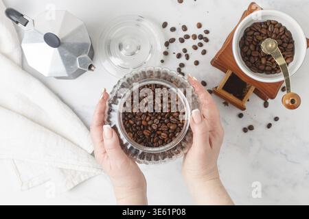 Eine Frau hält ein Glas gerösteten Kaffee in den Händen. Eine Kaffeekanne und eine manuelle Kaffeemühle auf einem Marmortisch. Selektiver Fokus Stockfoto