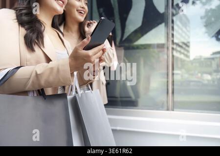 Eine Nahaufnahme zweier Freunde, die lächeln, während sie Einkaufstaschen und ein Smartphone in der Hand halten und die Aufregung des Black Friday festhalten Stockfoto